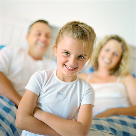 Girl folding arms and smiling in front of parents in bed, portrait Stock Photo - Premium Royalty-Free, Code: 695-05774227