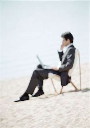 Businessman sitting on beach with laptop on knees, full length, close-up, blurred Stock Photo - Premium Royalty-Free, Code: 695-05776265