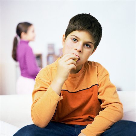 sister fights brother - Boy sitting on sofa, eating, girl standing in background Stock Photo - Premium Royalty-Free, Code: 695-05777046