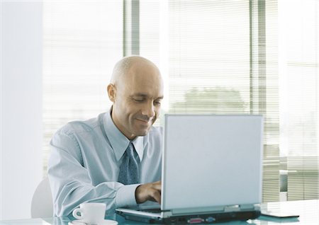 Businessman using laptop, smiling, coffee cup beside him on table Stock Photo - Premium Royalty-Free, Code: 695-05777546