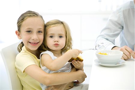 simsearch:695-03380035,k - Young girl sitting on her older sister's lap, holding spoon and croissant, smiling at camera Foto de stock - Royalty Free Premium, Número: 695-05779454