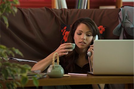 Young woman sitting at coffee table using phone and laptop computer, holding cup of tea Stock Photo - Premium Royalty-Free, Code: 695-05779675