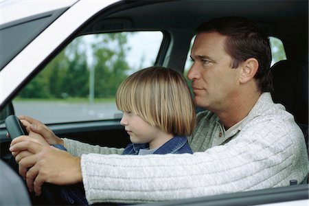 dad kid drive car - Man driving car with young son sitting on lap, both holding steering wheel Stock Photo - Premium Royalty-Free, Code: 695-05770106