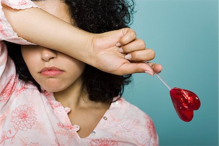 Woman frowning and covering eyes with arm, holding heart-shaped lollipop Stock Photo - Premium Royalty-Free, Code: 695-05770305