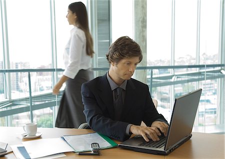 decision maker - Businessman using laptop, woman walking by in background Foto de stock - Sin royalties Premium, Código: 695-03374215