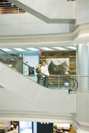 retail escalator - Couple taking escalator in shopping mall, waving at camera, in mid distance Stock Photo - Premium Royalty-Free, Code: 695-03374985