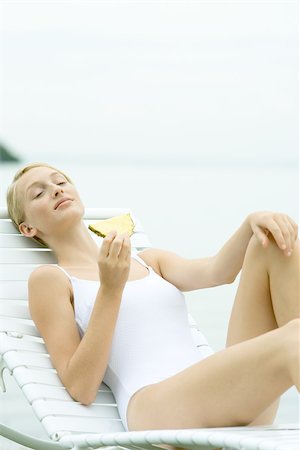 Teenage girl wearing bathing suit, sitting in lounge chair, holding slice of pineapple Stock Photo - Premium Royalty-Free, Code: 695-03375422