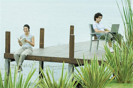 Young man and woman sitting apart on dock, woman reading book, man using laptop Stock Photo - Premium Royalty-Free, Code: 695-03376073