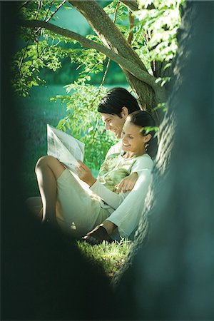 Couple sitting on ground, leaning against tree, reading newspaper, smiling, blurred tree trunk in foreground Stock Photo - Premium Royalty-Free, Code: 695-03376087