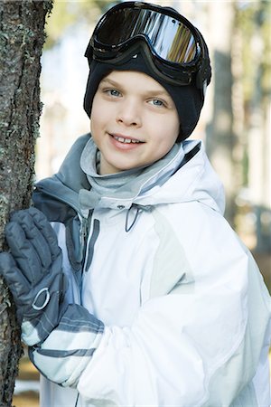 Boy wearing ski gear, leaning against tree trunk, smiling at camera, portrait Stock Photo - Premium Royalty-Free, Code: 695-03376426