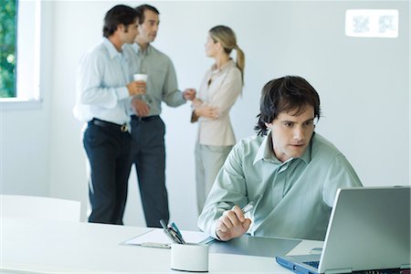 Businessman working on laptop, colleagues standing in background Stock Photo - Premium Royalty-Free, Code: 695-03376537