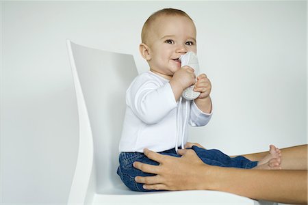 Baby sitting in chair putting shoe in mouth, mother's arms holding baby's legs Stock Photo - Premium Royalty-Free, Code: 695-03376617