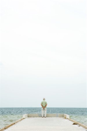 Senior man standing at end of pier, facing ocean, in distance, rear view Stock Photo - Premium Royalty-Free, Code: 695-03377561