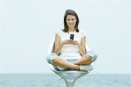 easy - Teenage girl sitting in chair outdoors, holding cell phone, smiling at camera Stock Photo - Premium Royalty-Free, Code: 695-03377914
