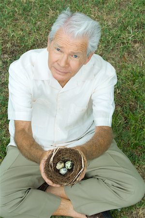 Senior man sitting on the ground, holding bird's nest with eggs, looking up at camera Stock Photo - Premium Royalty-Free, Code: 695-03377910