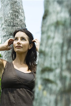 simsearch:695-03378068,k - Woman leaning against tree trunk, pushing hair behind ears, looking up Fotografie stock - Premium Royalty-Free, Codice: 695-03378062