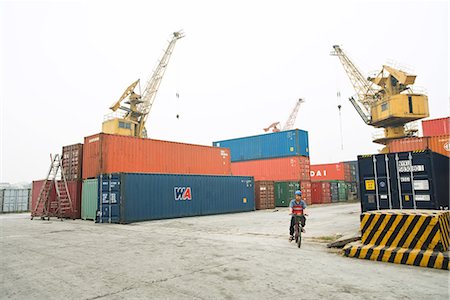 simsearch:614-06974220,k - Person riding bicycle past metal storage units, cranes in background Stock Photo - Premium Royalty-Free, Code: 695-03378130