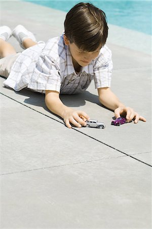 Boy lying on ground, playing with toy cars, next to pool Stock Photo - Premium Royalty-Free, Code: 695-03378178