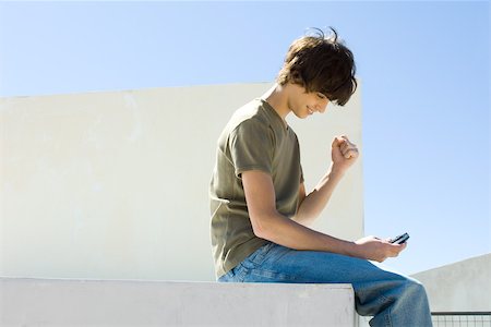 Teenage boy sitting outdoors, looking down at cell phone, making a fist, smiling Stock Photo - Premium Royalty-Free, Code: 695-03378589