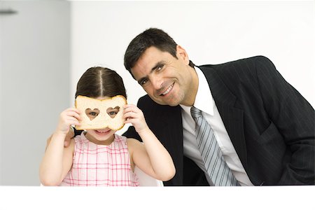 Father and daughter sitting side by side, girl looking through slice of bread with heart-shaped holes, both smiling at camera Stock Photo - Premium Royalty-Free, Code: 695-03378614