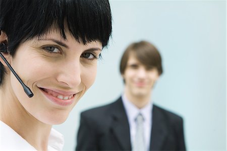 simsearch:632-03779793,k - Woman wearing headset, smiling at camera, businessman in background Foto de stock - Royalty Free Premium, Número: 695-03378935