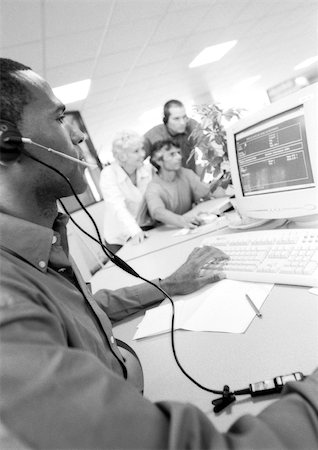 Man with headset using computer, colleagues in blurred background, B&W Stock Photo - Premium Royalty-Free, Code: 695-03384079