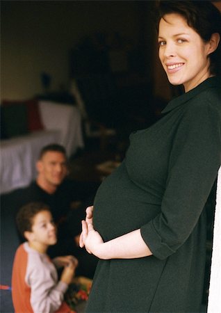 simsearch:695-03384752,k - Pregnant woman standing and holding stomach, man and child sitting in background Foto de stock - Royalty Free Premium, Número: 695-03384763