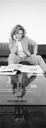 Woman sitting on sofa with newspaper open on coffee table, looking at camera, b&w, vertical Stock Photo - Premium Royalty-Free, Code: 695-03385603