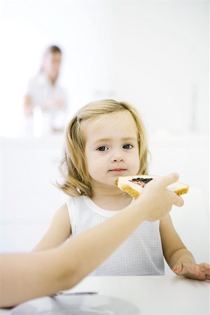 simsearch:695-03380035,k - Young girl sitting at table, being fed a piece of bread with jam, cropped view Foto de stock - Royalty Free Premium, Número: 695-03389994