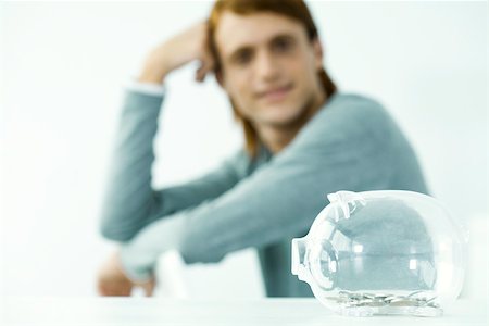 pig bank - Transparent piggy bank sitting on table, young man in background holding head Stock Photo - Premium Royalty-Free, Code: 695-03380301