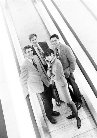 simsearch:695-03382908,k - Group of business people on moving walkway smiling at camera, business woman in front on cell phone, b&w. Fotografie stock - Premium Royalty-Free, Codice: 695-03382912