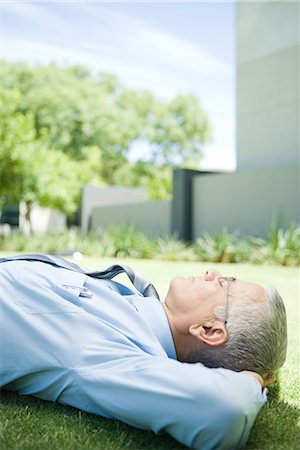 Mature businessman lying on ground outdoors, hands behind head, eyes closed, side view Stock Photo - Premium Royalty-Free, Code: 696-03394326