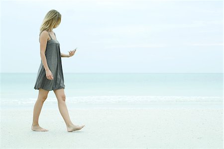 Young woman walking on the beach, looking at cell phone Stock Photo - Premium Royalty-Free, Code: 696-03394641