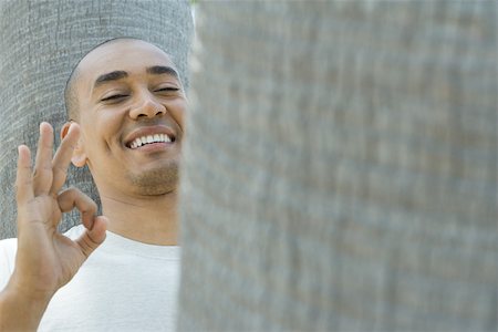 simsearch:633-02417937,k - Man leaning against tree trunk, making OK sign, smiling at camera Stock Photo - Premium Royalty-Free, Code: 696-03395231