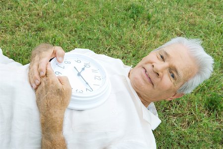 simsearch:695-03377910,k - Senior man lying on grass, holding clock, smiling at camera Fotografie stock - Premium Royalty-Free, Codice: 696-03395256