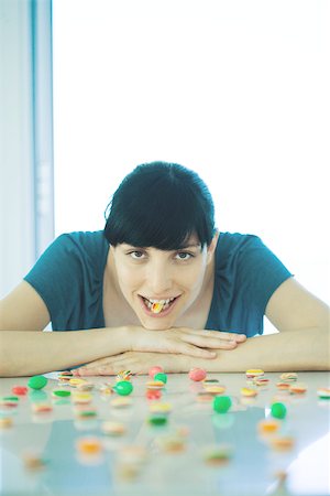 simsearch:695-03375576,k - Young woman sitting at table covered with candy, holding piece of candy between teeth Foto de stock - Sin royalties Premium, Código: 696-03401454