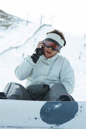 Young snowboarder sitting on the ground, using cell phone, looking at camera Stock Photo - Premium Royalty-Free, Code: 696-03401840