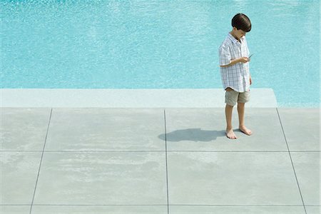 Boy standing by swimming pool, looking at cell phone, high angle view Stock Photo - Premium Royalty-Free, Code: 696-03401975