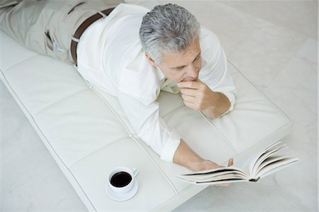 Mature man lying on stomach on chaise longue, reading book, cup of coffee nearby, high angle view Stock Photo - Premium Royalty-Free, Code: 696-03402021