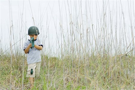 Little boy wearing toy helmet and grenade, pointing toy machine gun at camera, standing in tall grass Stock Photo - Premium Royalty-Free, Code: 696-03402033