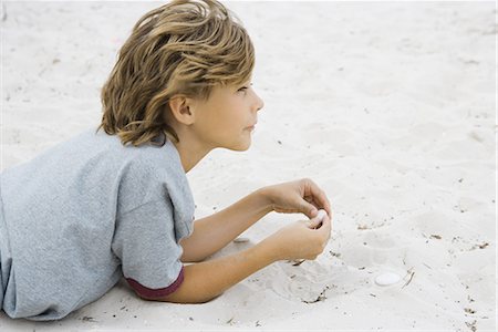 Boy lying on sand, holding pebble, looking away, side view Stock Photo - Premium Royalty-Free, Code: 696-03402043