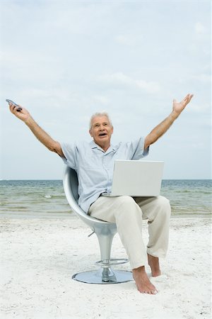 Senior man sitting on beach with laptop on lap, arms raised, shouting Stock Photo - Premium Royalty-Free, Code: 696-03402147