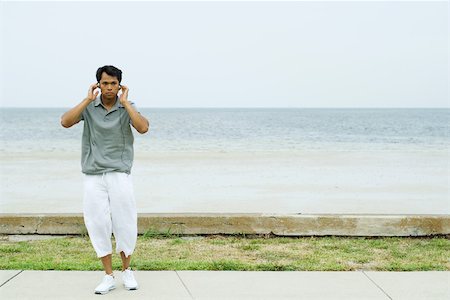 Man standing in front of beach, listening to headphones, hands raised to head Stock Photo - Premium Royalty-Free, Code: 696-03402265