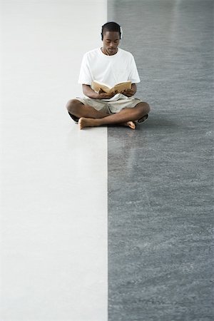 Teenage boy sitting cross-legged on the floor, reading a book, listening to headphones Stock Photo - Premium Royalty-Free, Code: 696-03402475