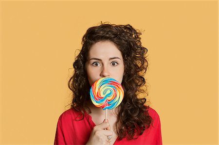 Portrait of a young woman holding lollipop in front of face over colored background Stock Photo - Premium Royalty-Free, Code: 693-06121347