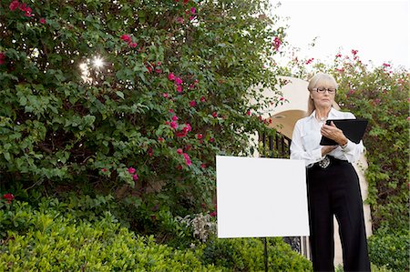 Senior real estate agent standing by sign board reading from clipboard Stock Photo - Premium Royalty-Free, Code: 693-06324009