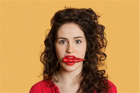 Portrait of a young woman with red chili pepper in mouth over colored background Stock Photo - Premium Royalty-Free, Code: 693-06324107