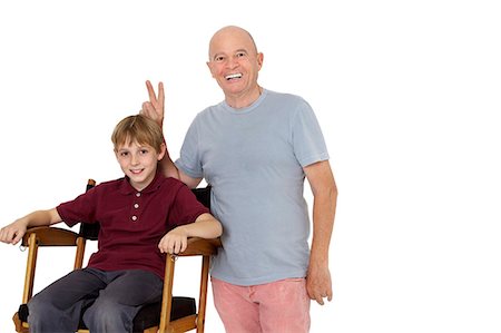 Portrait of senior man gesturing peace sign while pre-teen boy sitting on director's chair over white background Stock Photo - Premium Royalty-Free, Code: 693-06324815