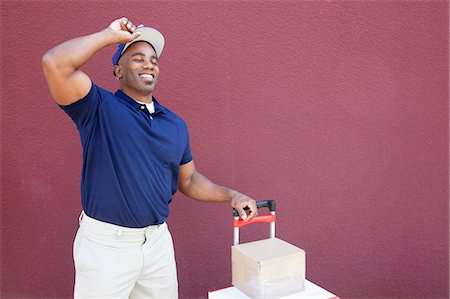 Happy young African American delivery man standing with handtruck over colored background Stock Photo - Premium Royalty-Free, Code: 693-06323984