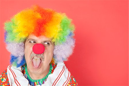 Senior male clown sticking out tongue while looking away over red background Fotografie stock - Premium Royalty-Free, Codice: 693-06378847